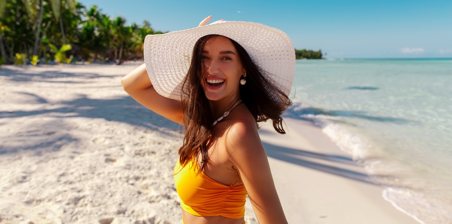 Beautiful happy lady wearing straw hat at beach with turquoise ocean on background, looking at camera and laughing, enjoying tropical vacation